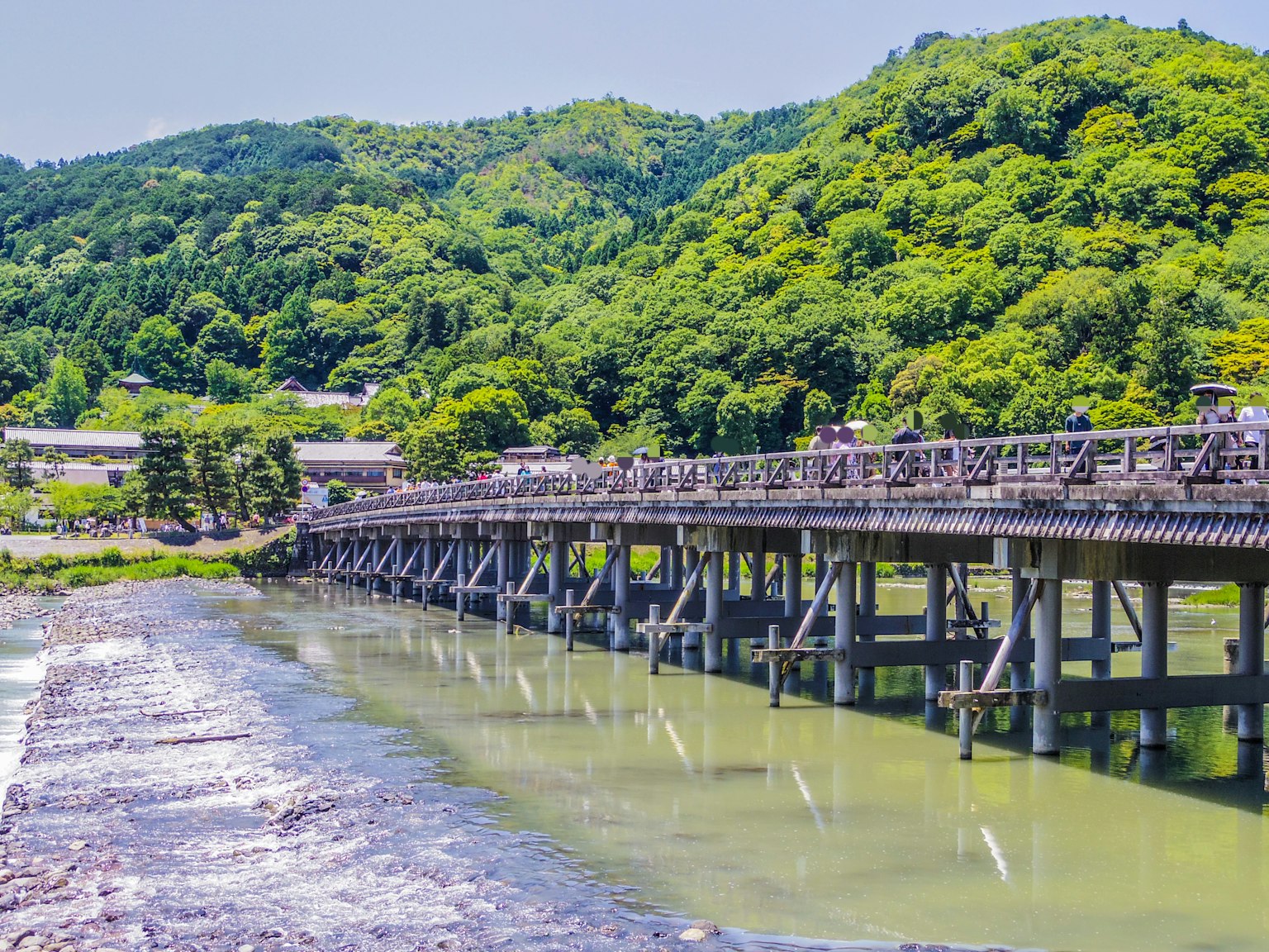 Jembatan kayu yang melintasi sungai dikelilingi bukit hijau
