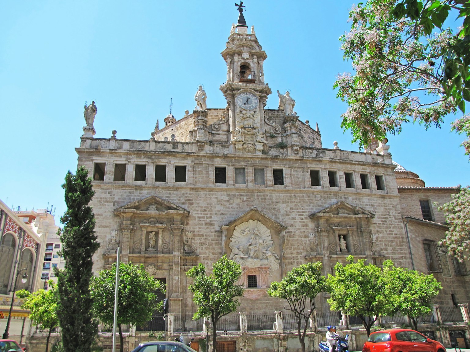 Edificio grandioso en Valencia con hermosa fachada de piedra bajo un cielo azul con árboles verdes y coches rojos