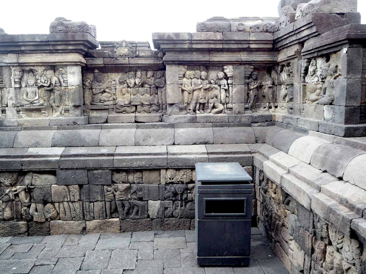 Mur en pierre avec des sculptures anciennes et une poubelle noire