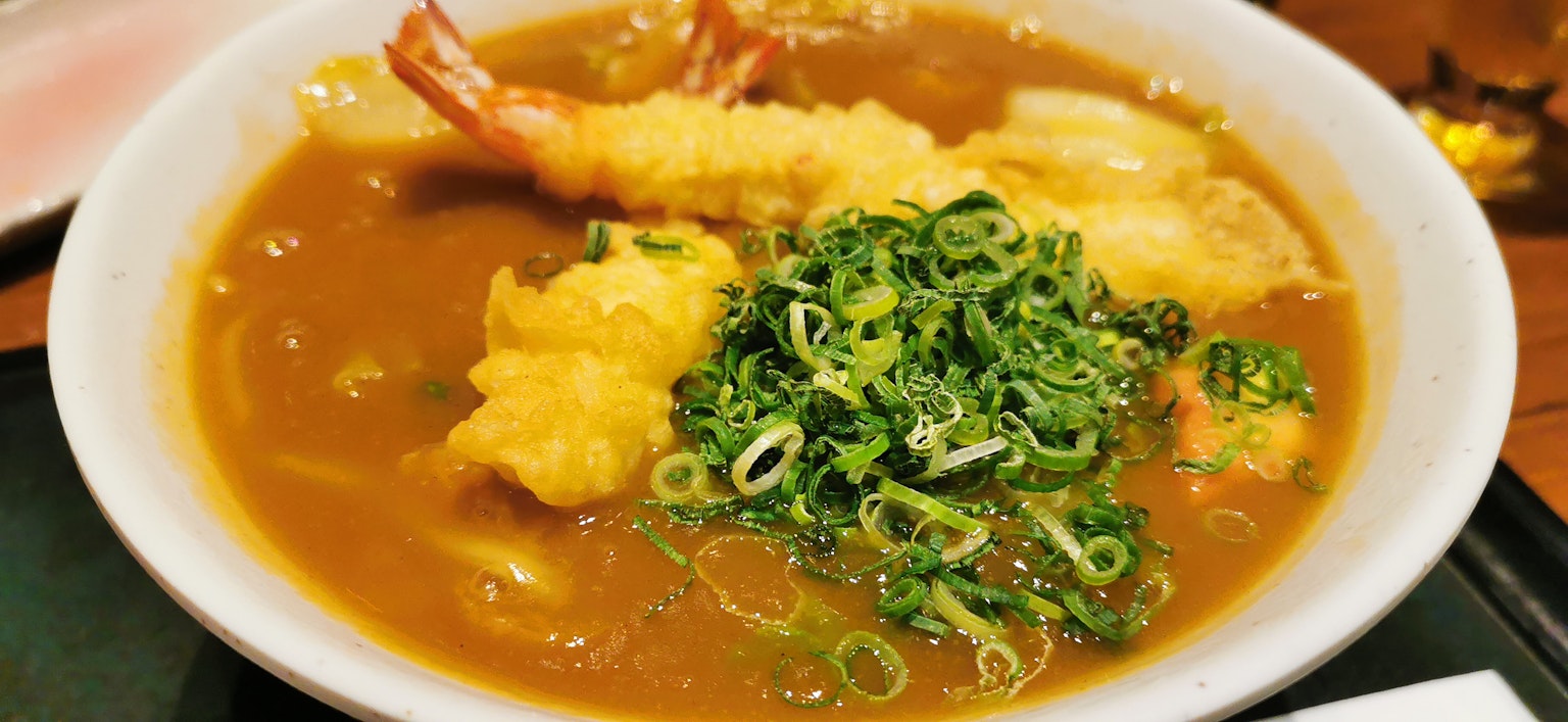 Ciotola di udon al curry guarnita con gamberi tempura e cipollotti