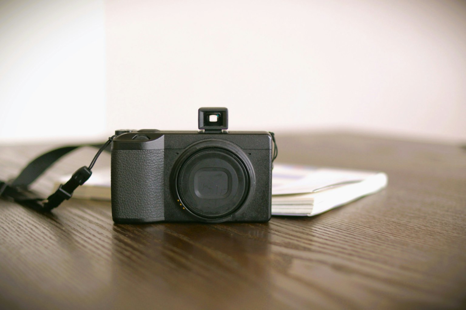 Una cámara digital colocada sobre una mesa de madera con un cuaderno cerca La cámara es negra con un lente visible