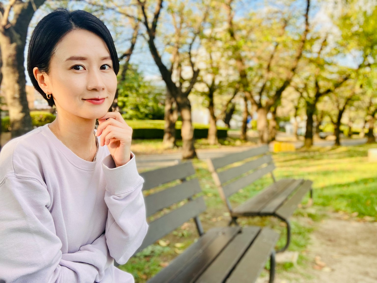 Potret seorang wanita yang duduk di bangku taman dengan ekspresi tenang dan pohon sakura di latar belakang