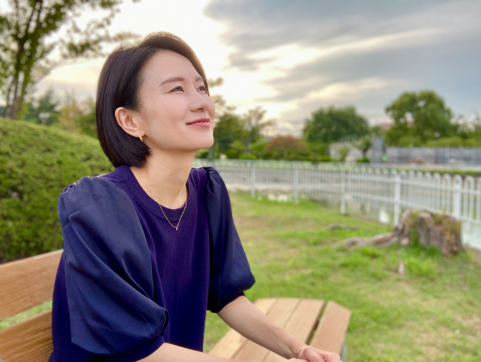 Người phụ nữ mỉm cười khi ngồi trên ghế công viên với bầu trời xanh và cây cối