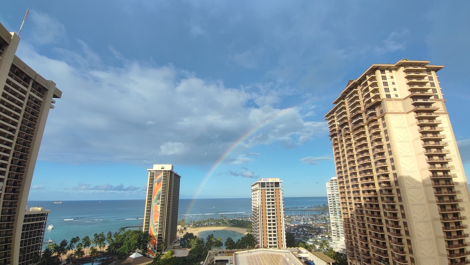 A scenic view of the ocean in Hawaii with blue skies and a rainbow high-rise buildings in the foreground