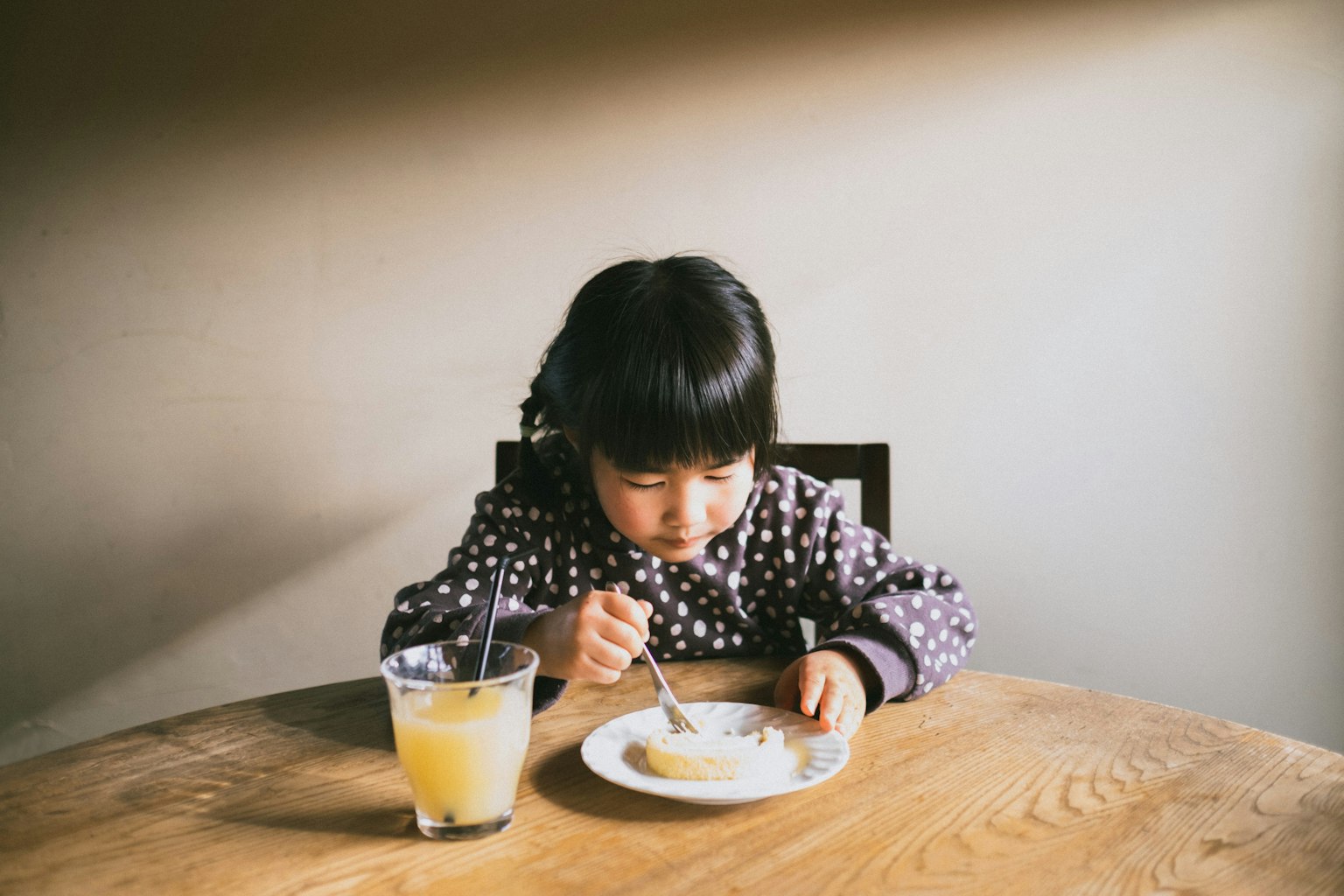 Child sitting at a table eating food from a plate glass of orange juice nearby