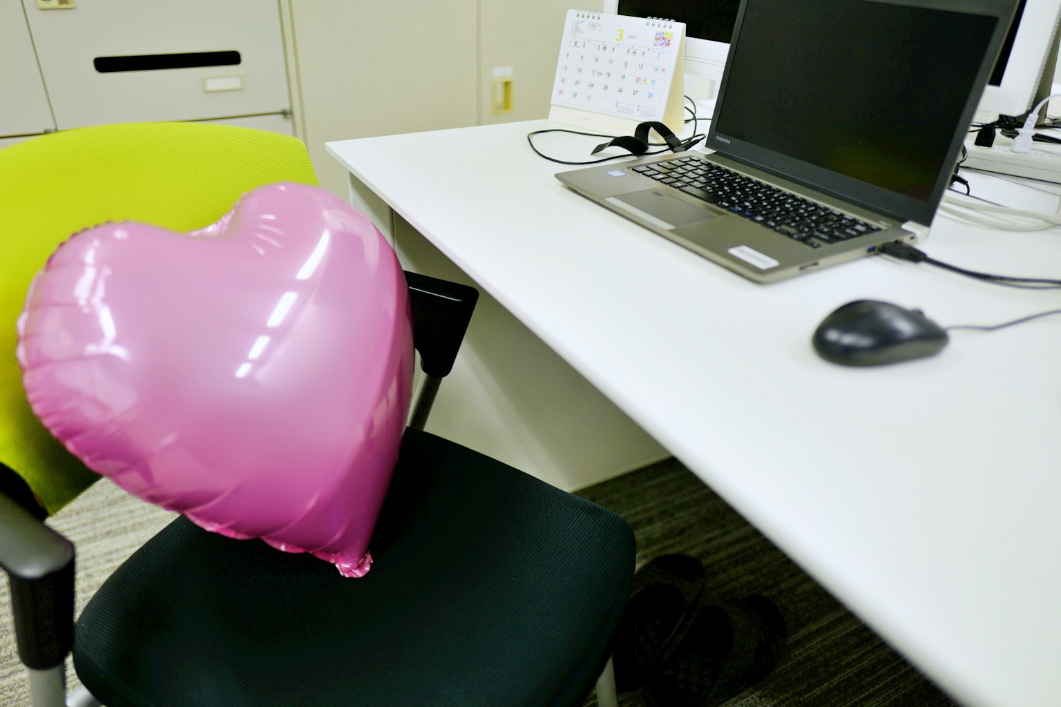 Palloncino a forma di cuore rosa appoggiato su una sedia verde accanto a una scrivania con computer e mouse