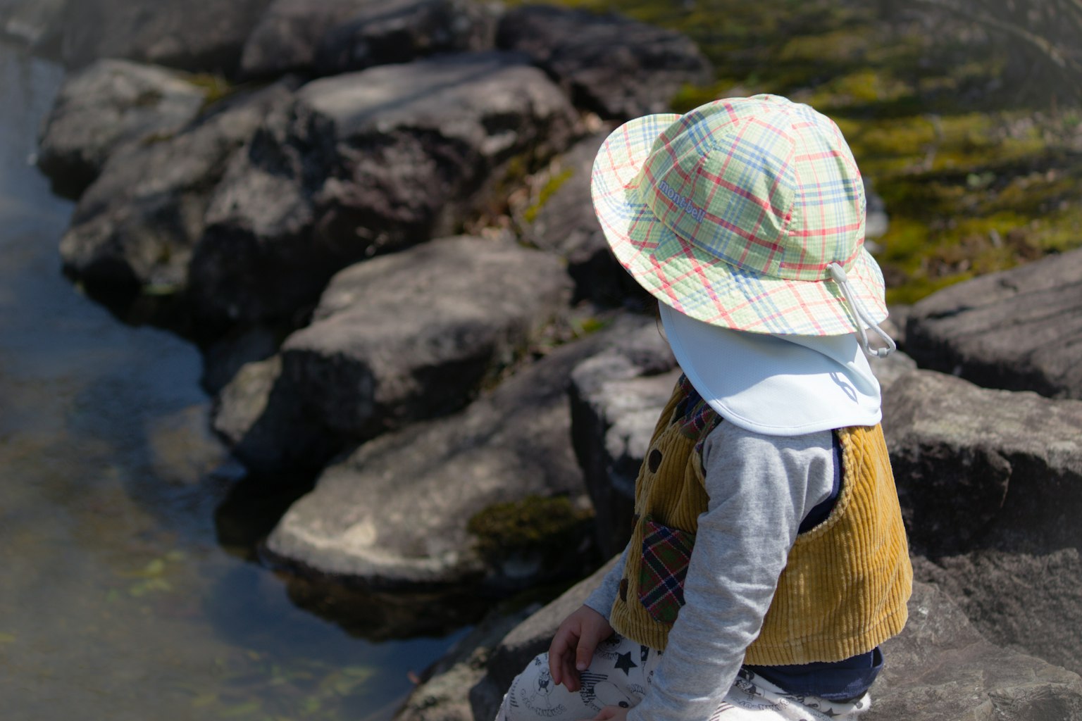 Ein Kind sitzt auf Felsen mit einem Hut und Weste in der Nähe von Wasser