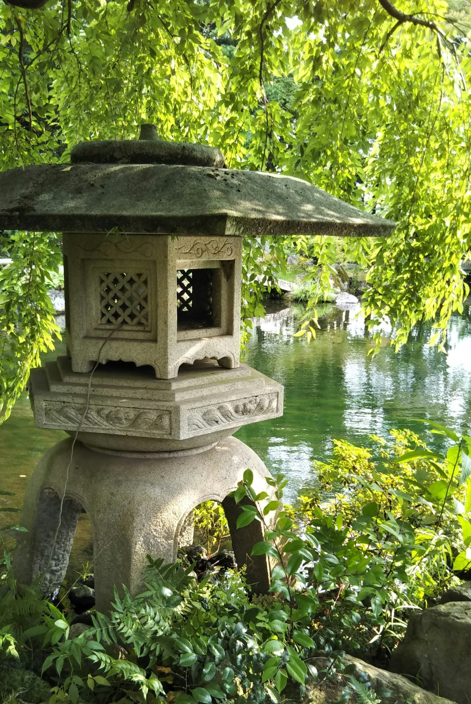 Eine Steinlaterne in einem japanischen Garten umgeben von üppigem Grün in der Nähe eines ruhigen Teiches