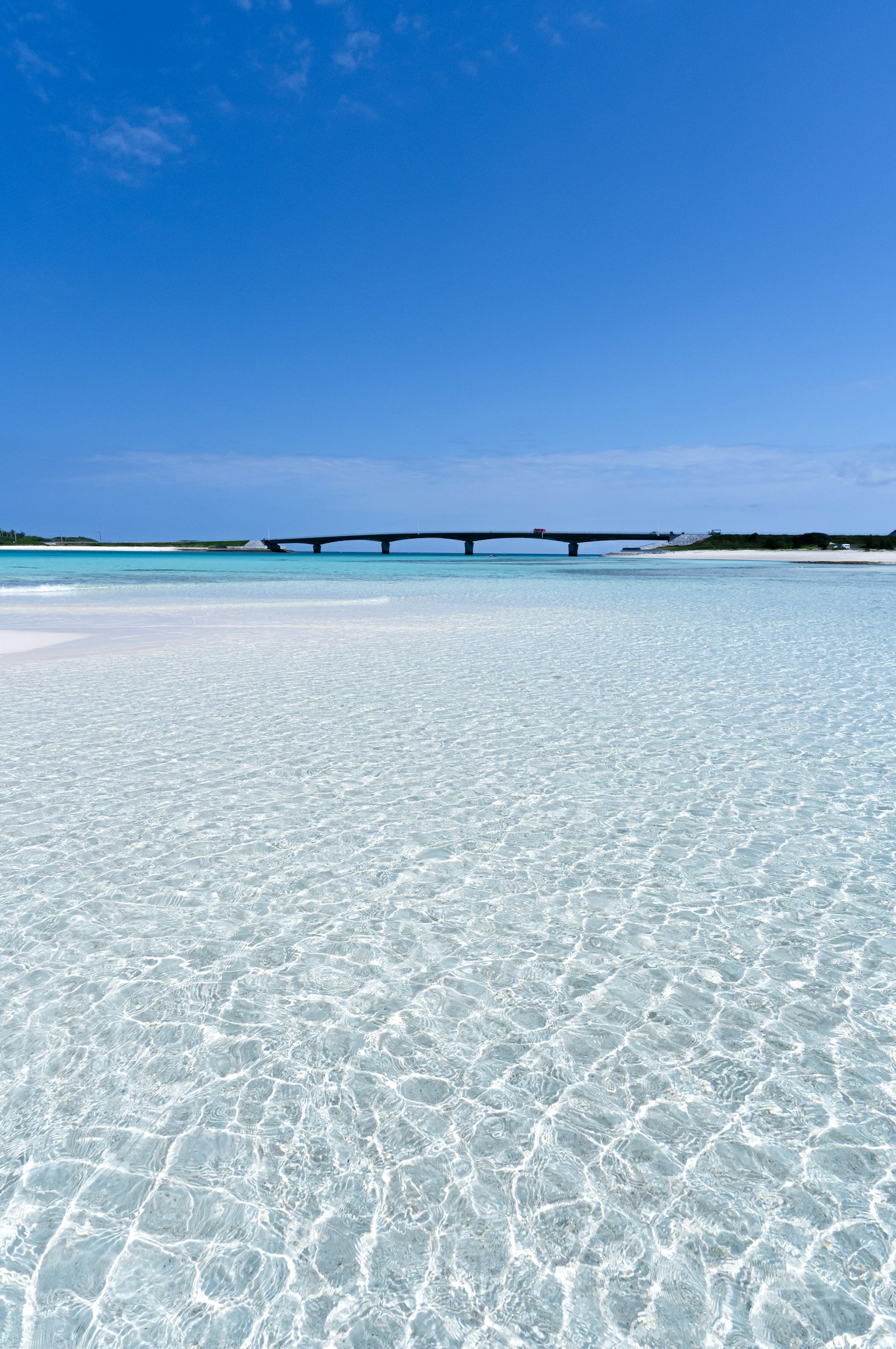 透明な水面と青い空が広がるビーチの風景 橋が遠くに見える