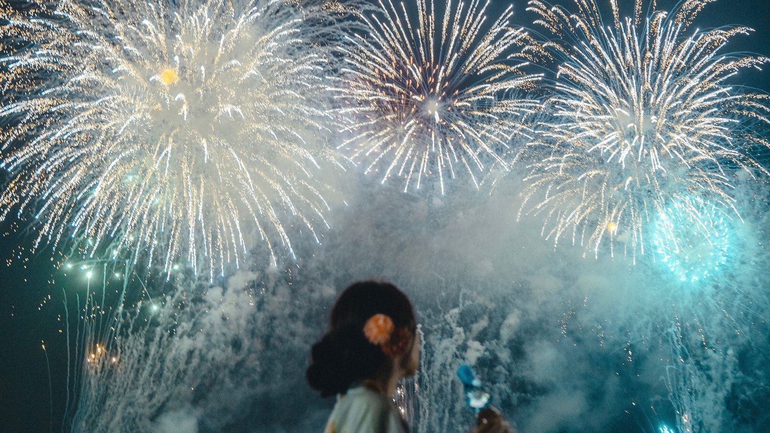 A woman holding a glass with fireworks bursting in the night sky