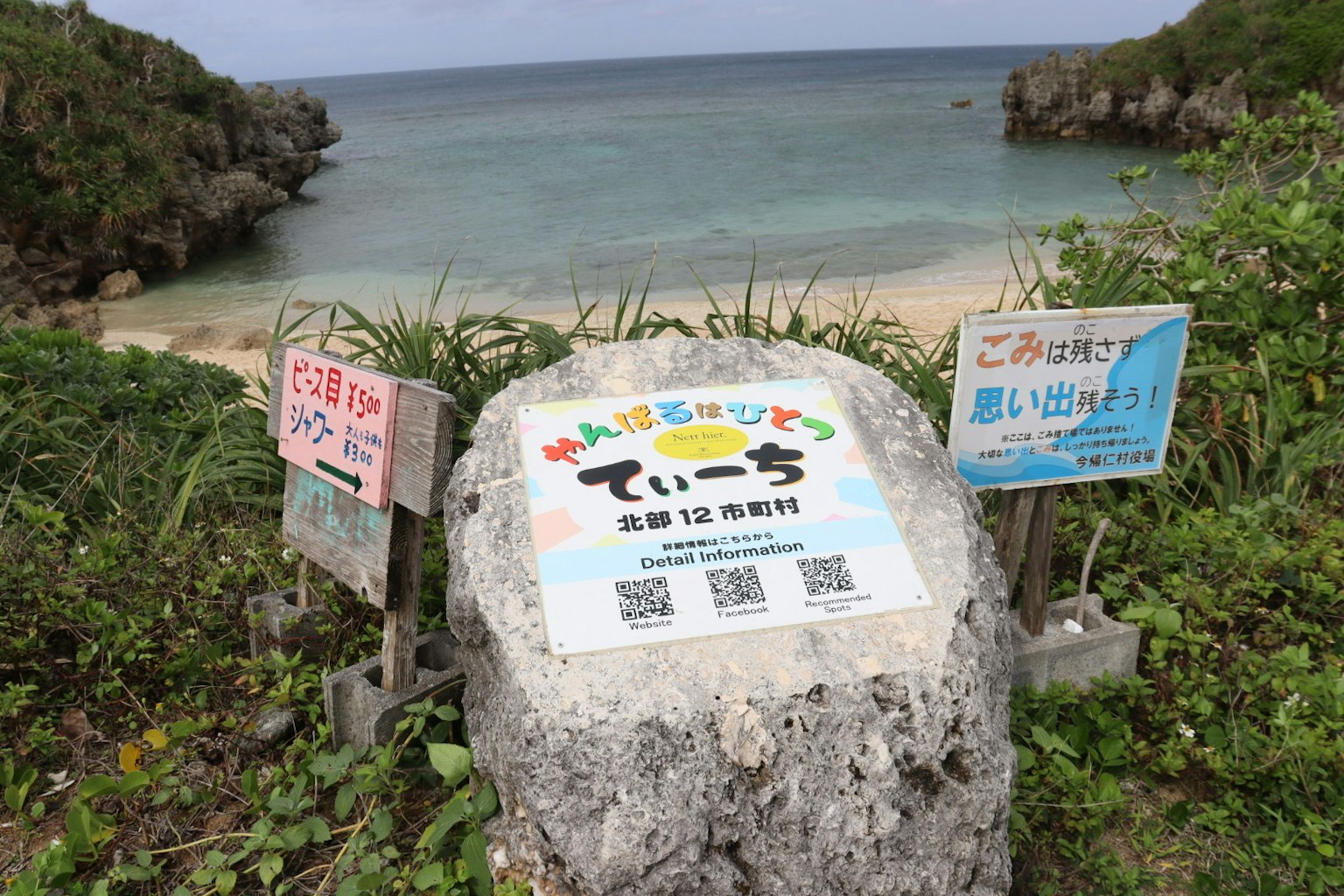 Señal de piedra que dice 'てぃーち' rodeada de hierba verde y mar azul