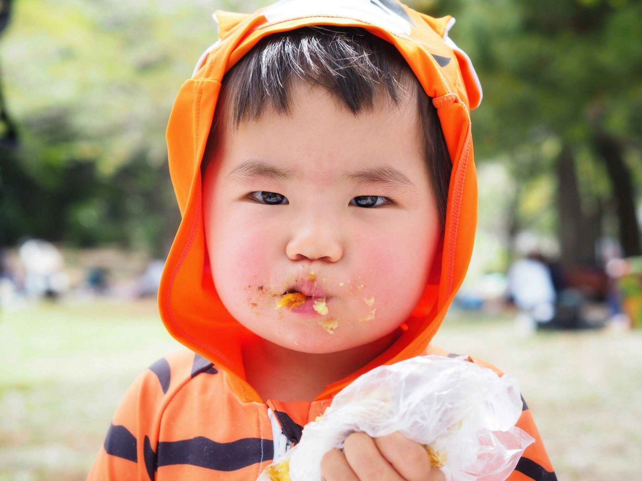 オレンジのフードをかぶった子どもが公園で食べ物をほおばる
