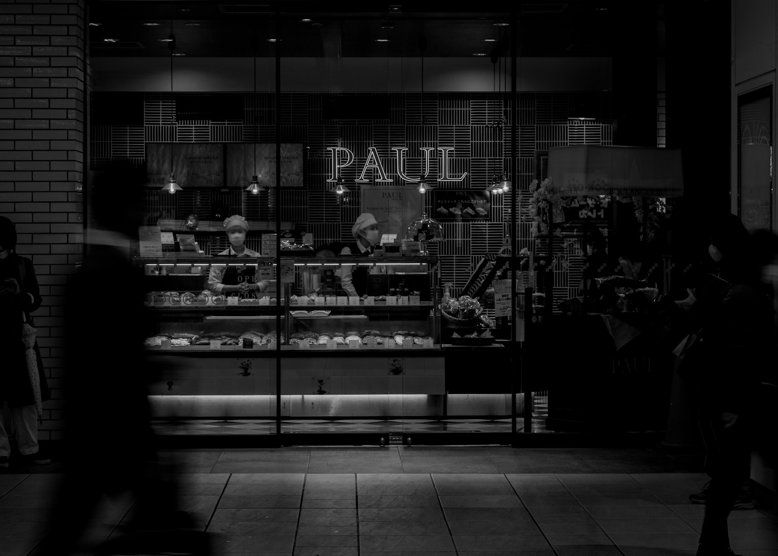 Bakery shop named PAUL illuminated at night with people passing by