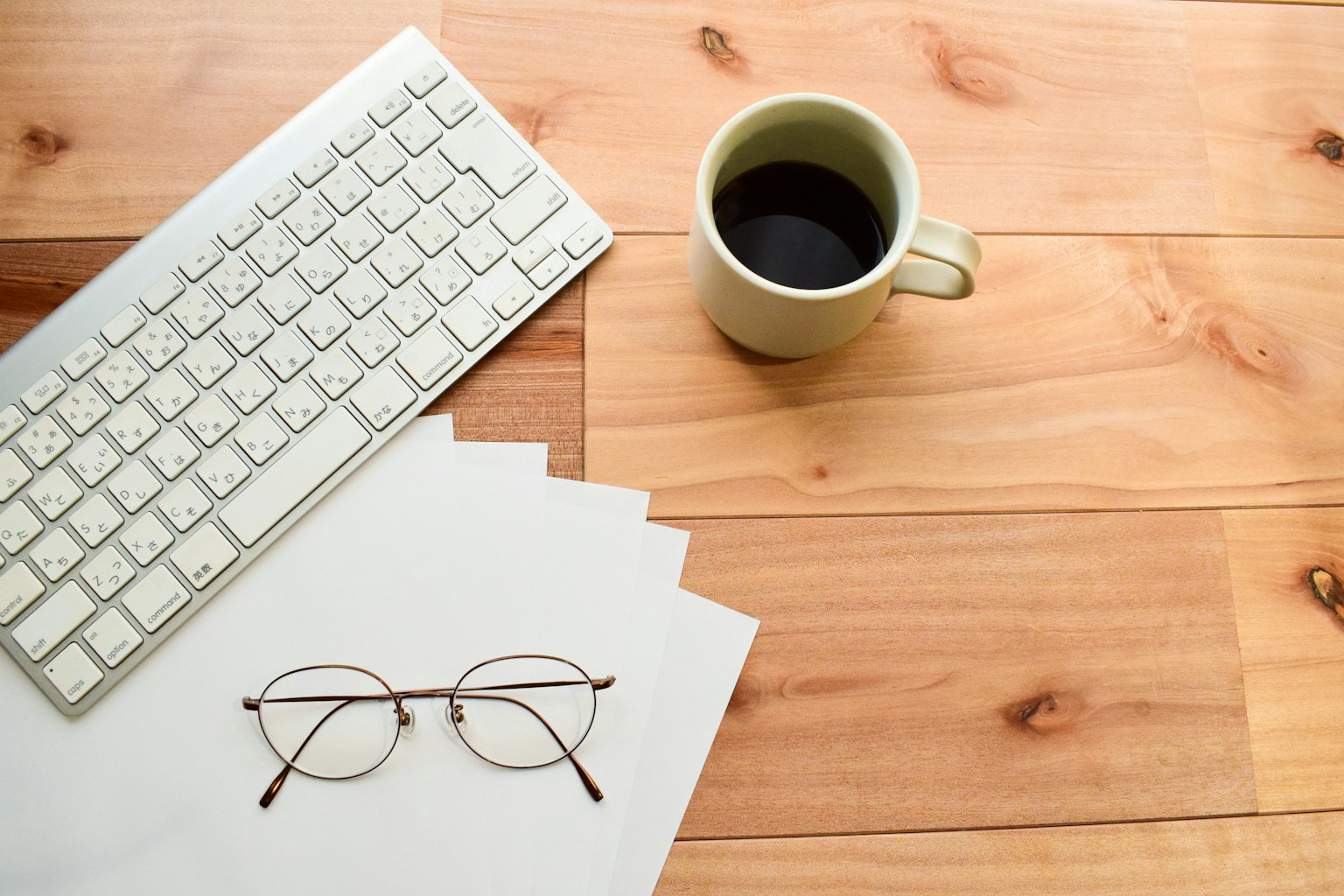 Weißes Keyboard und Kaffeetasse auf einem Holztisch mit Brille und Blättern Papier in der Nähe