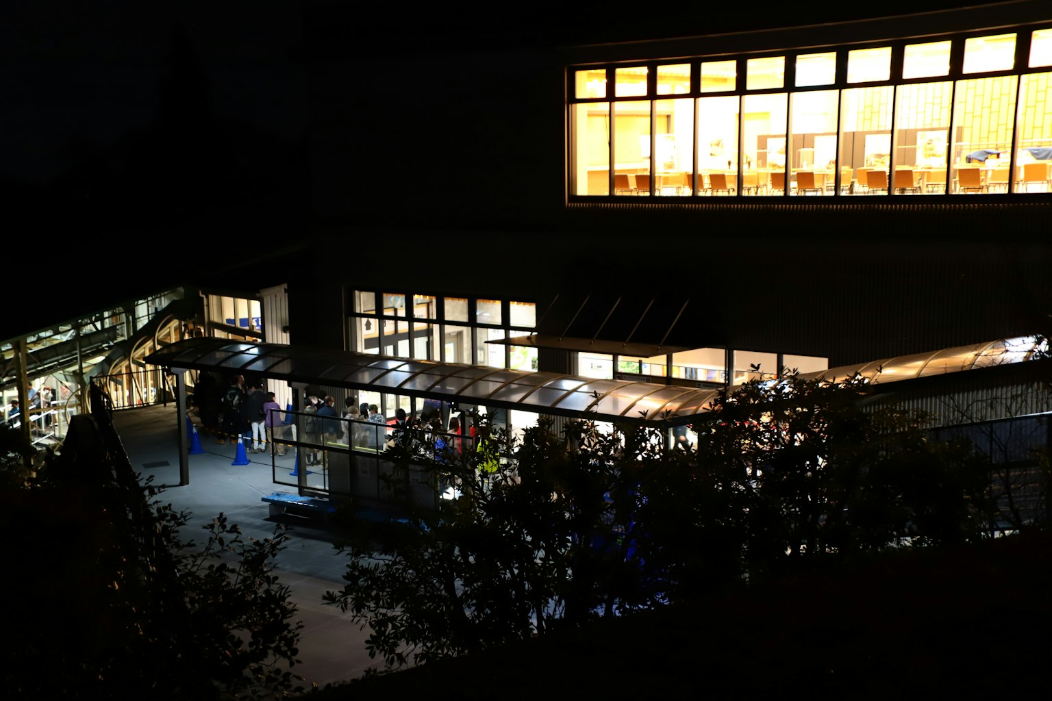 Eksterior perpustakaan di malam hari dengan orang-orang yang masuk dan jendela yang terang