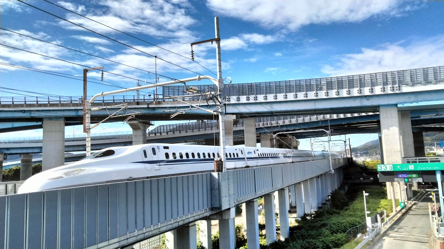 Tàu Shinkansen chạy dưới một cấu trúc cao với bầu trời xanh và dây điện