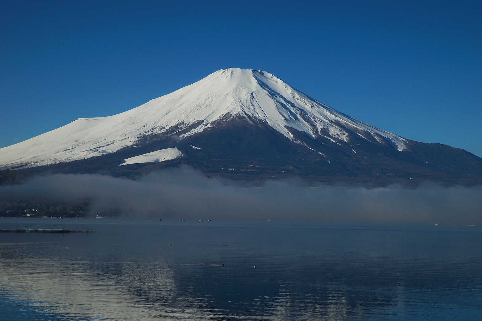 Monte Fuji con una cima innevata che si riflette in un lago calmo sotto un cielo blu chiaro