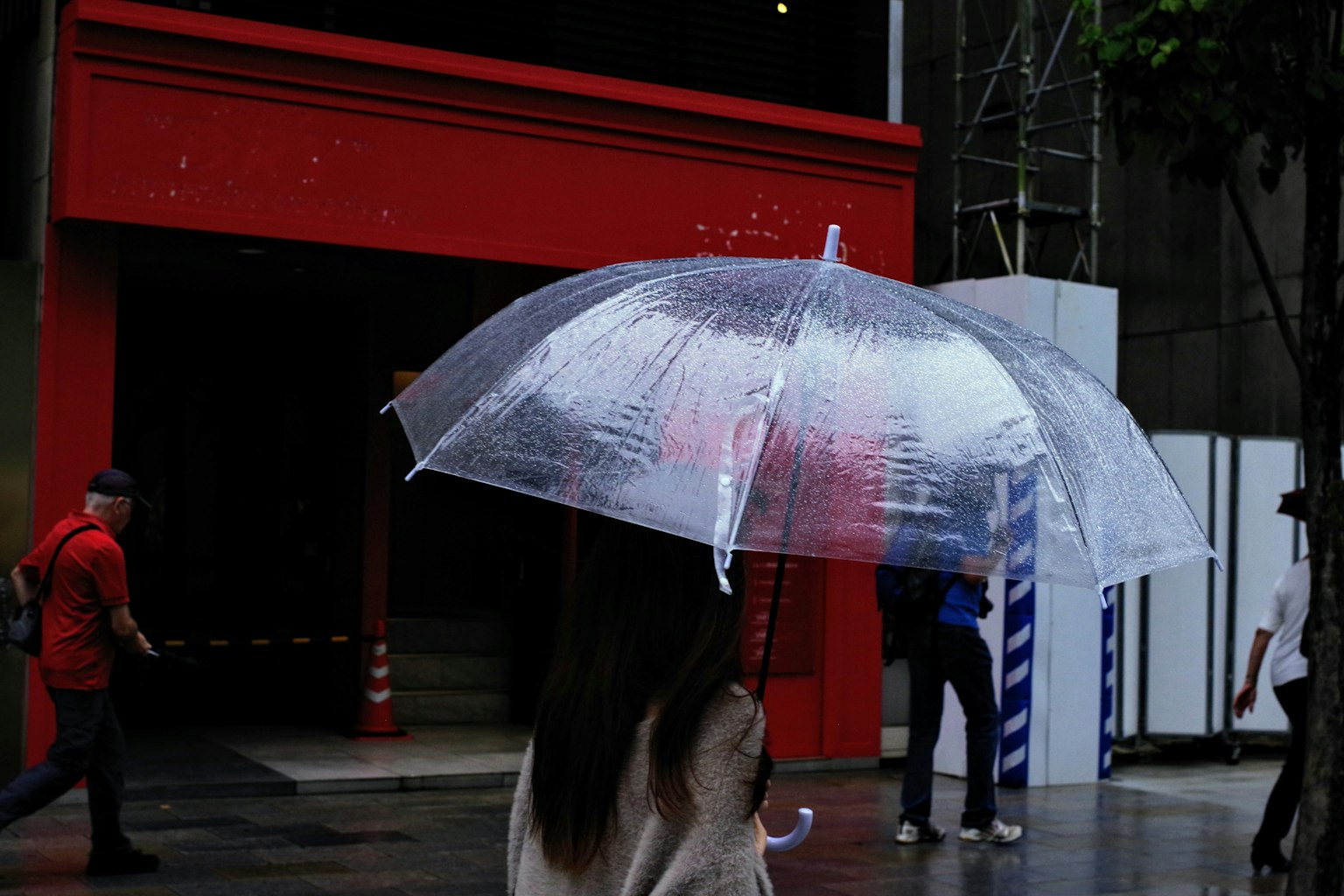 ผู้หญิงคนหนึ่งเดินถือร่มใสในฉากเมืองที่มีอาคารสีแดงอยู่เบื้องหลัง