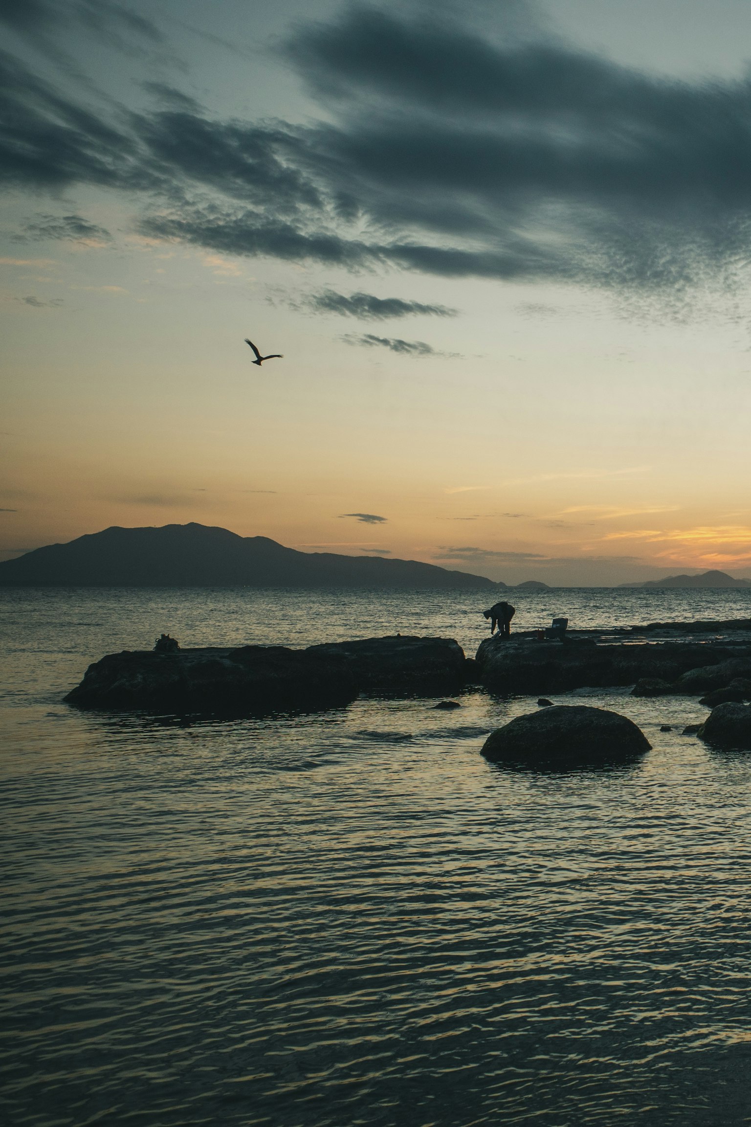 Una persona in piedi su rocce al mare durante il tramonto con montagne in lontananza