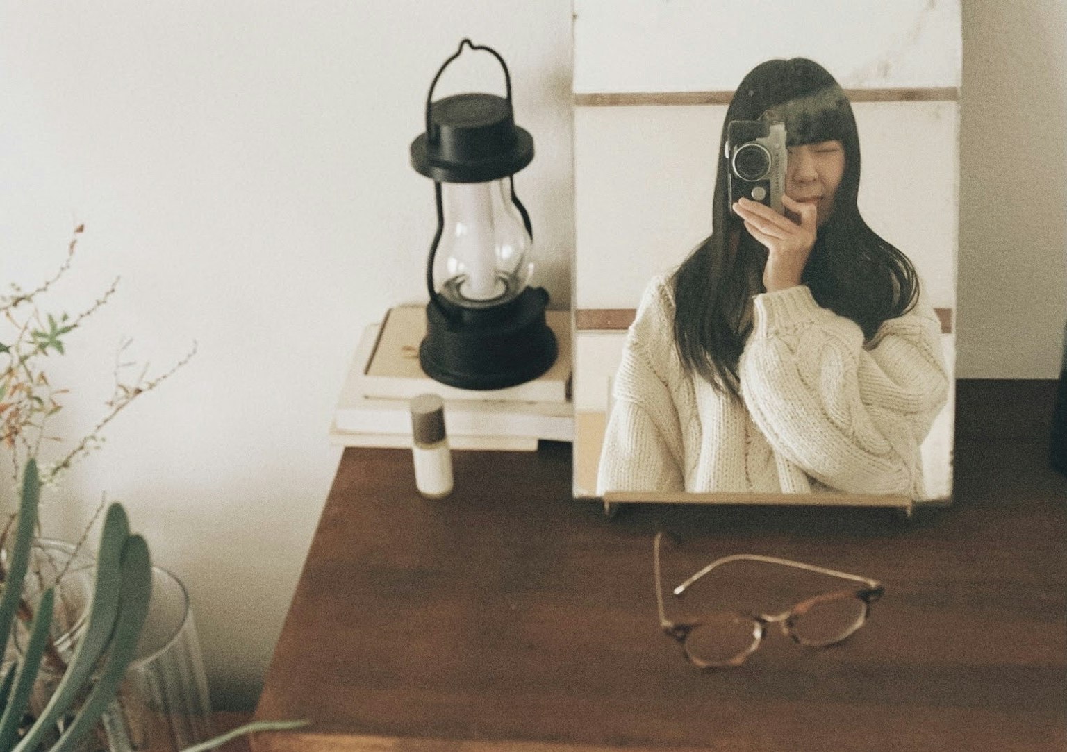 Une femme se regardant dans un miroir sur une table en bois avec une lanterne et des lunettes à proximité