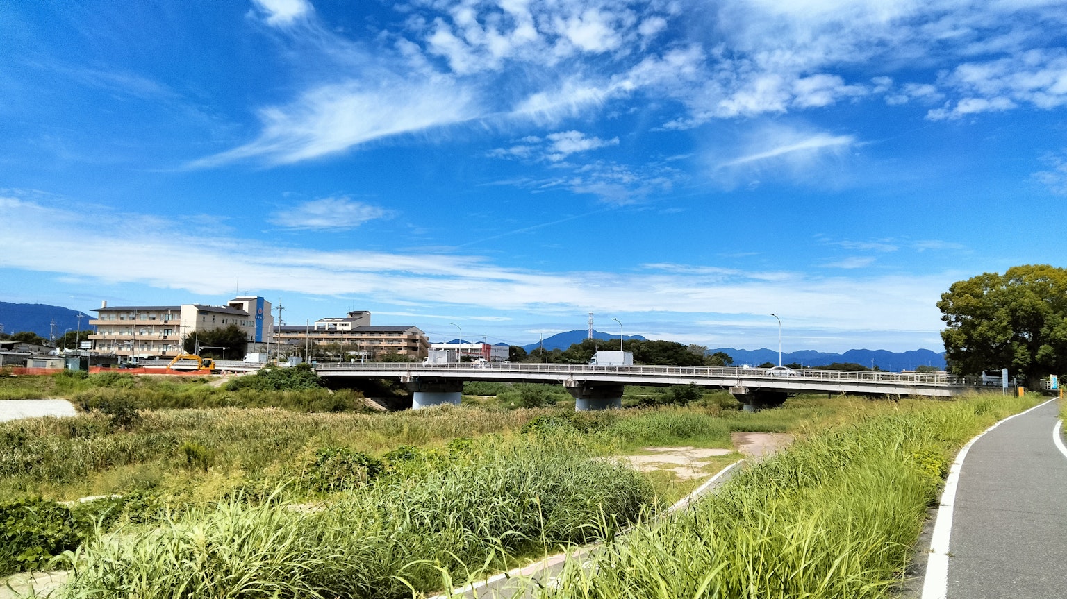 青空と雲を背景にした川沿いの風景 緑の草と道路が見える
