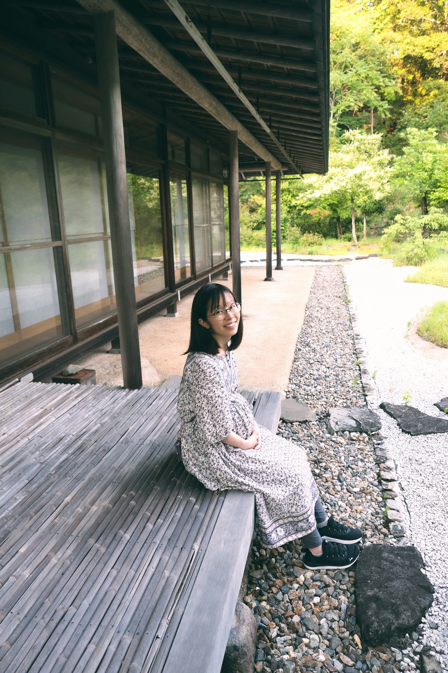 Wanita duduk di dek kayu dikelilingi oleh alam dan arsitektur tradisional