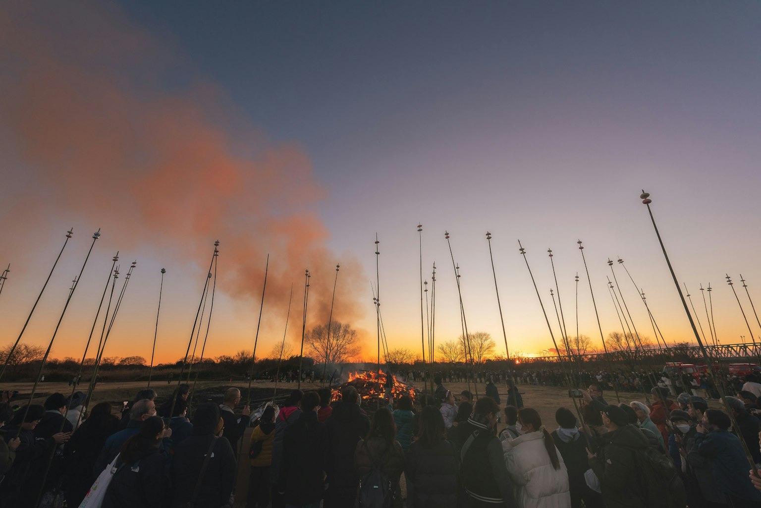 Folla che osserva il fumo sollevarsi al tramonto con pali in silhouette