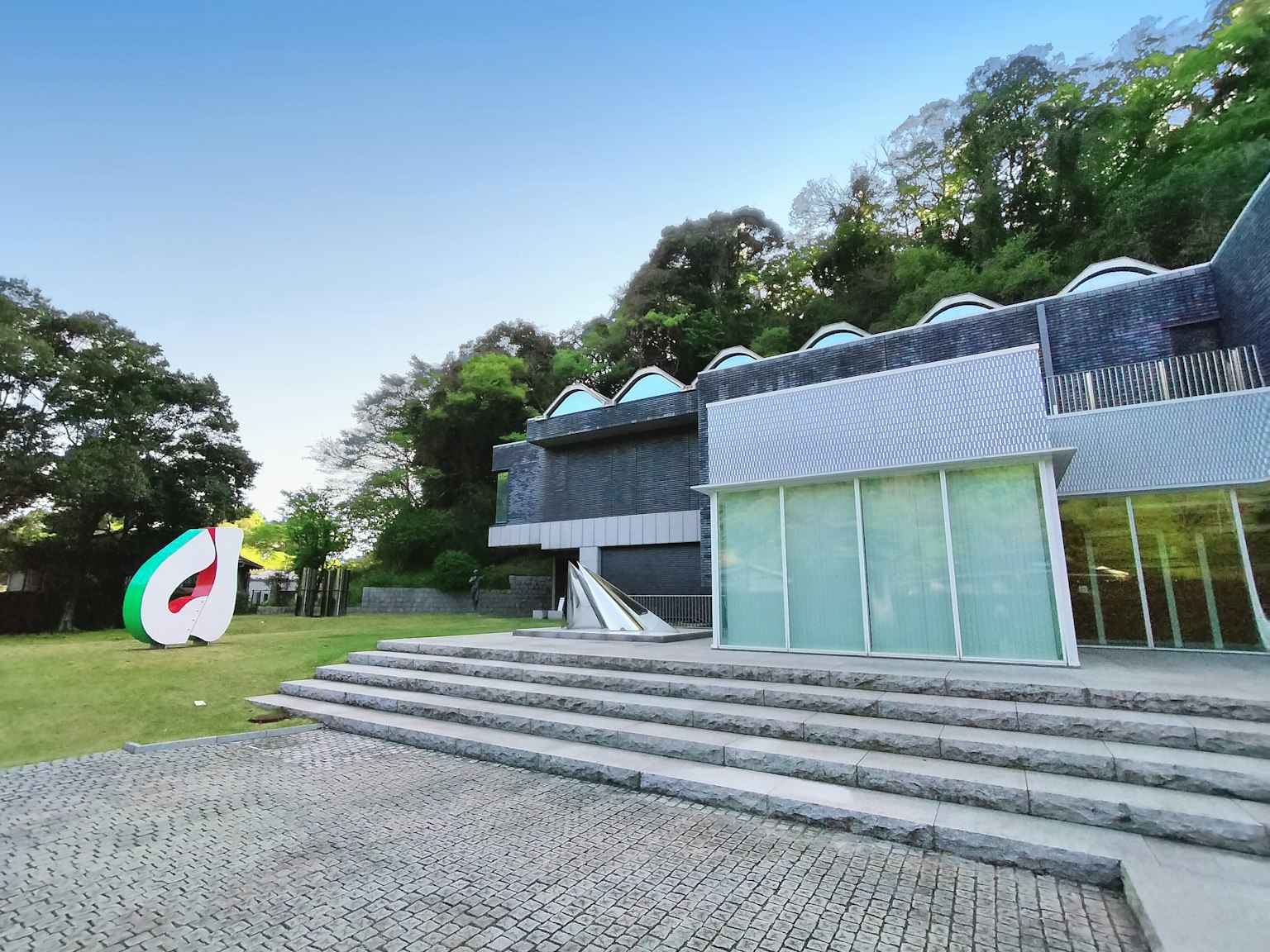 Bâtiment moderne avec une façade en verre et un toit unique entouré de verdure Sculpture colorée sur la pelouse