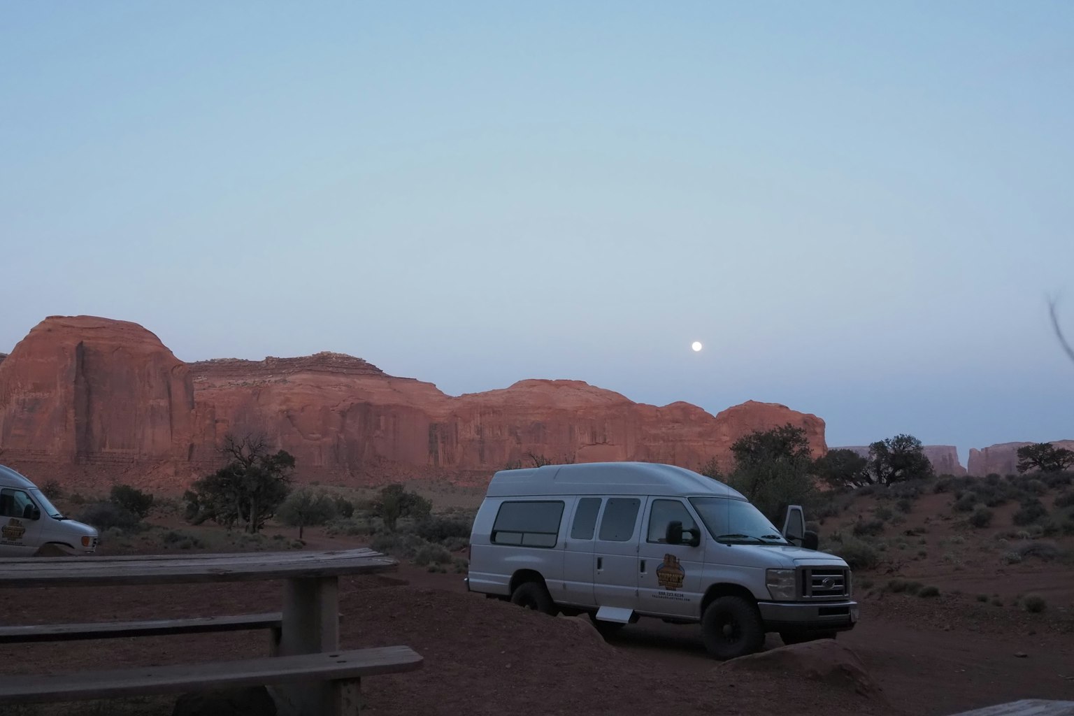 Furgón blanco estacionado en Monument Valley al anochecer con la luna al fondo