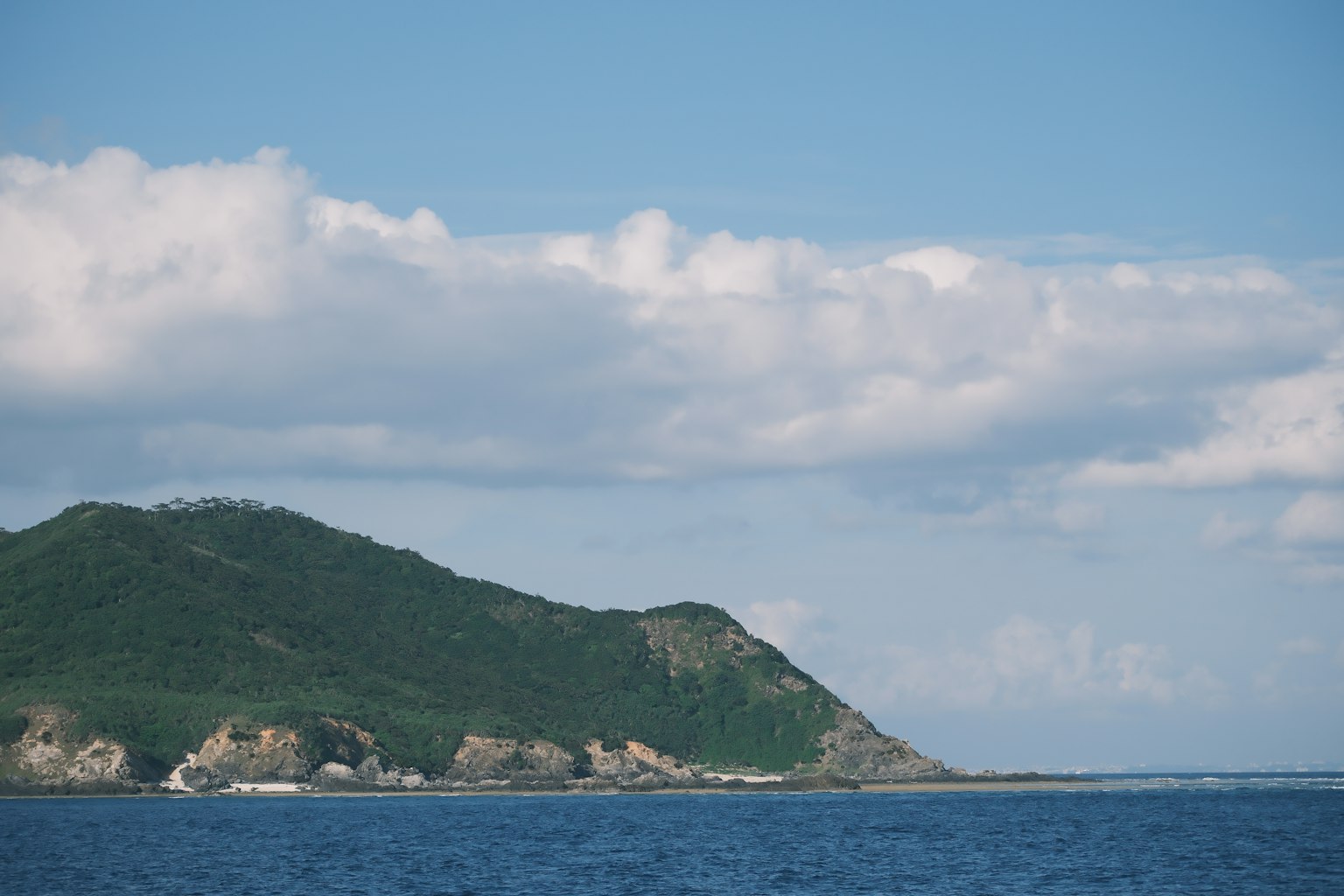 A green mountain rises above a blue sea under a sky with white clouds