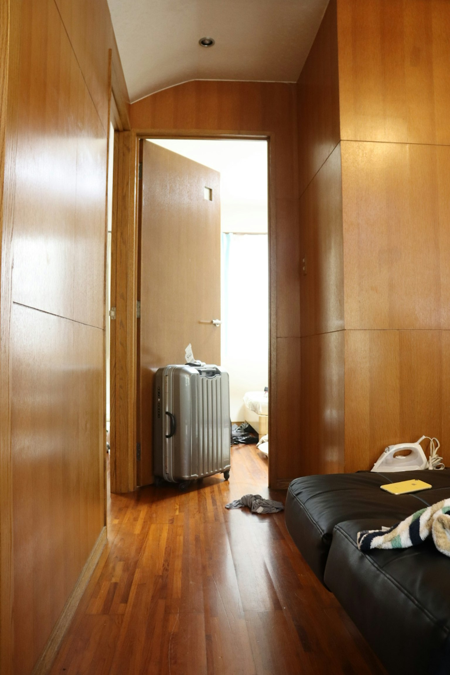 Image d'un couloir étroit avec des murs et un sol en bois Une valise est placée près d'une porte ouverte avec une lumière vive visible au-delà