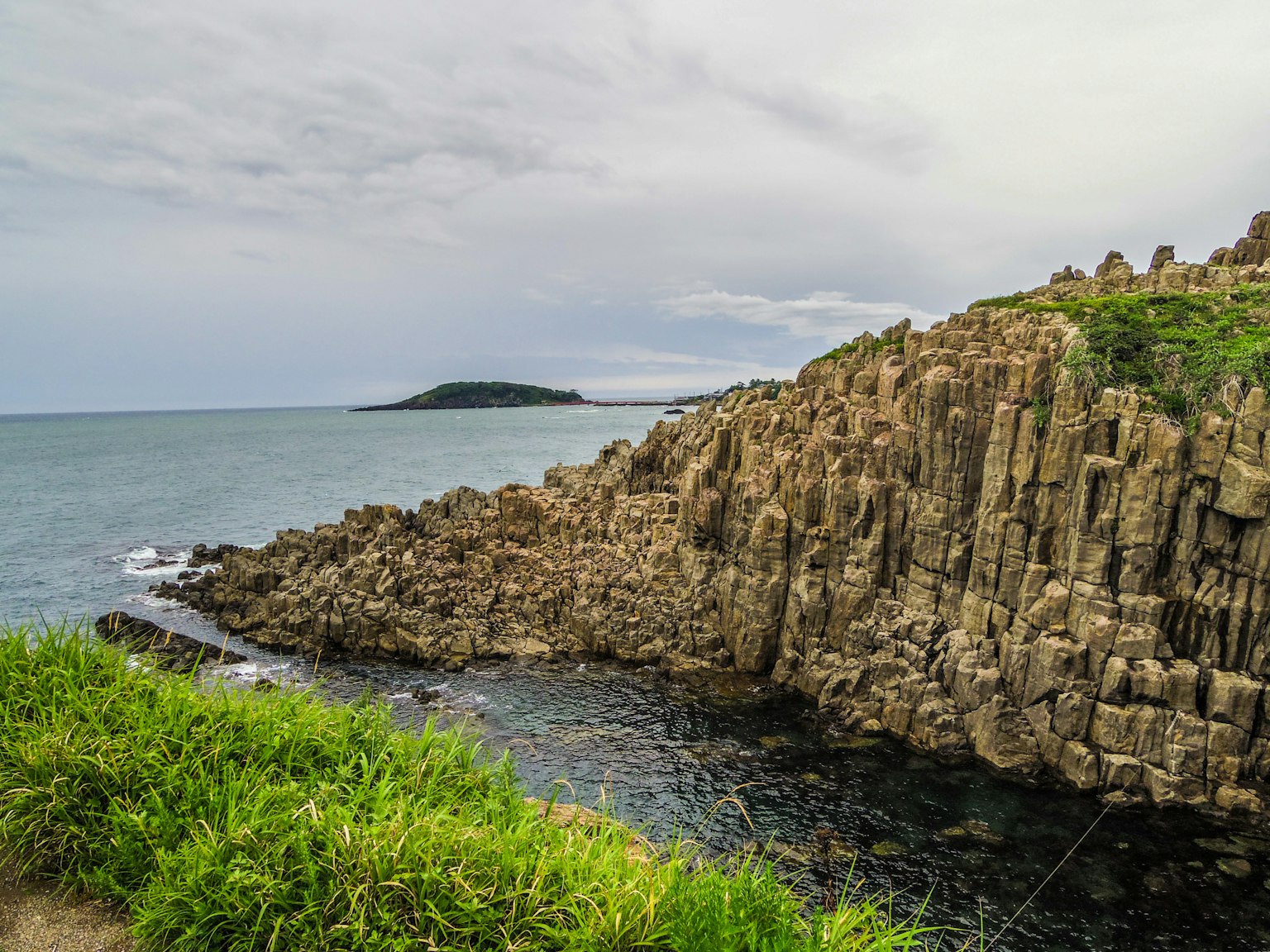 Pemandangan pantai dengan tebing berbatu dan tumbuhan hijau dengan pulau di kejauhan
