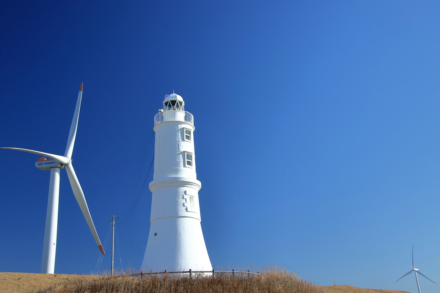 Weißer Leuchtturm neben Windkraftanlagen unter einem klaren blauen Himmel
