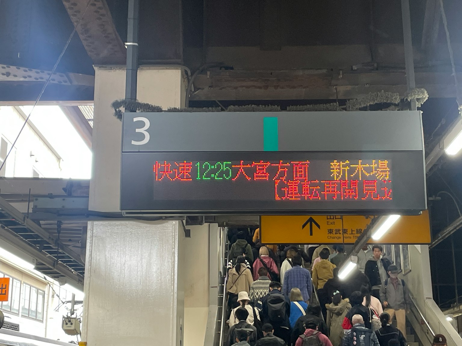 Digital display board at a train station showing train information with passengers on escalators