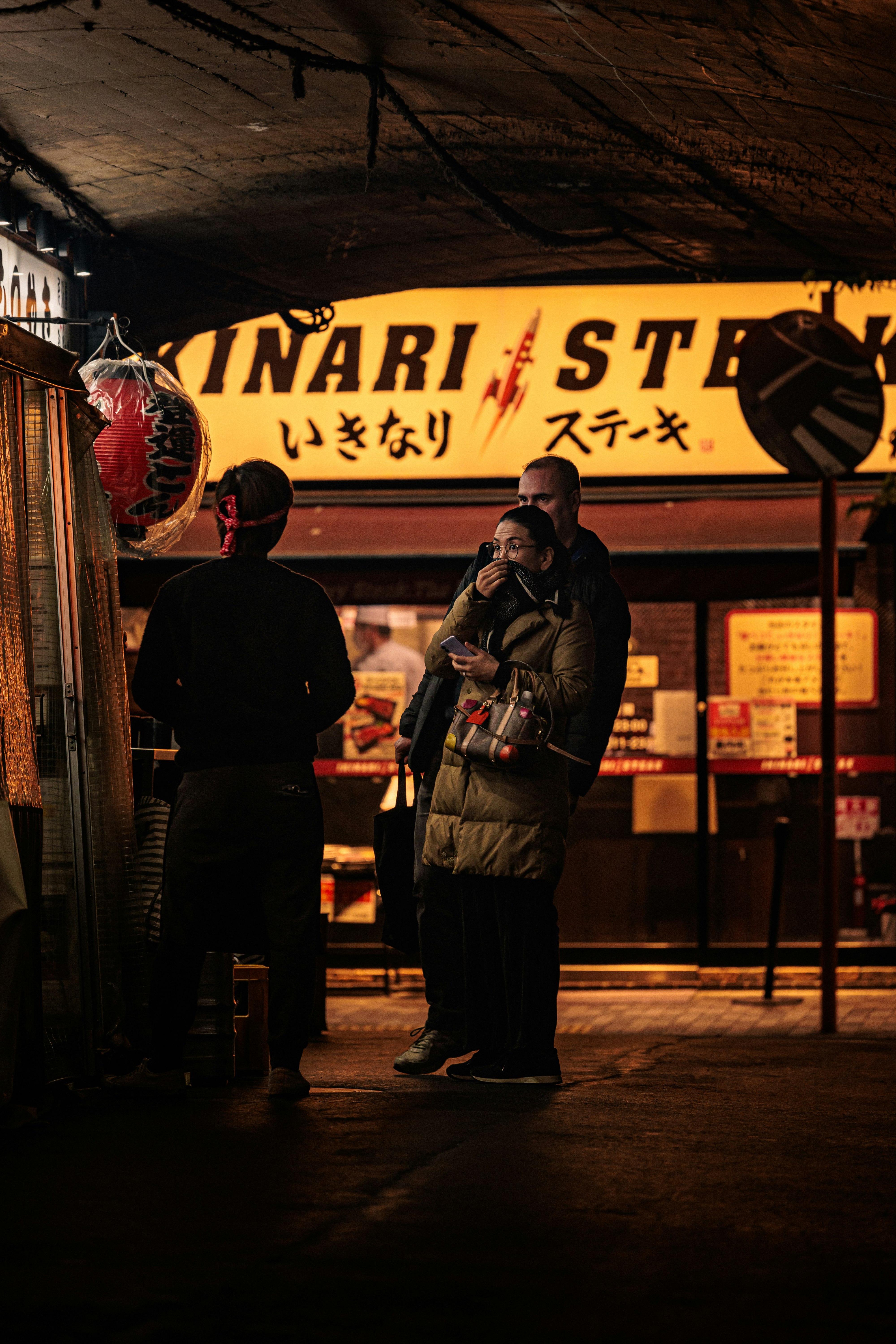 Scena notturna in strada fuori da un chiosco di « inari steak » con persone vicino all’insegna