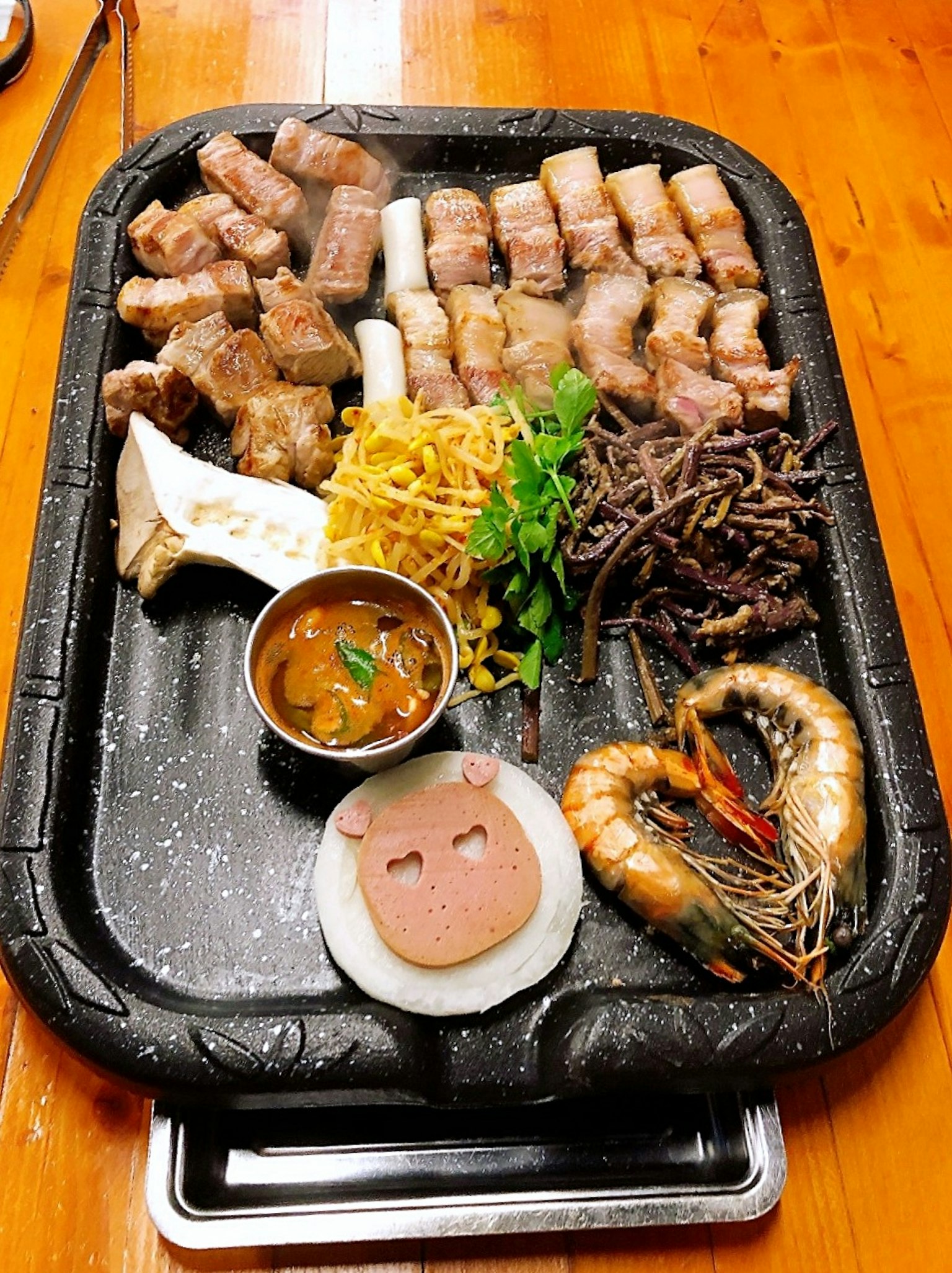 Piring daging panggang dengan udang, daging babi, sayuran, dan saus di atas nampan hitam