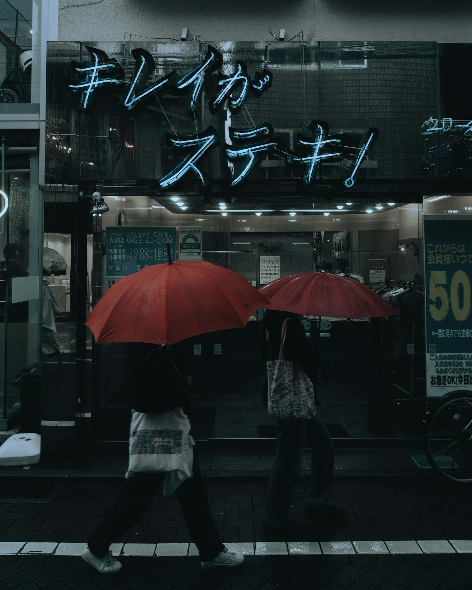 Two people walking in the rain with red umbrellas in front of a Japanese store sign