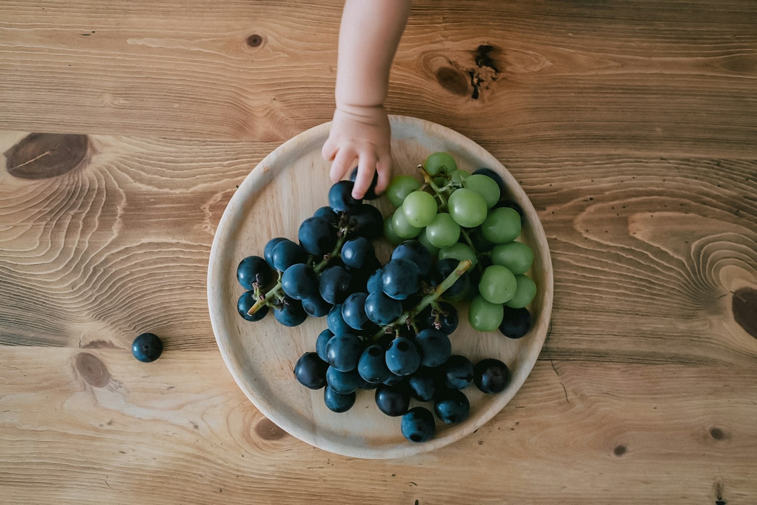 木製のテーブルの上にある青いブドウと緑のブドウの盛り合わせに手を伸ばす子供の手