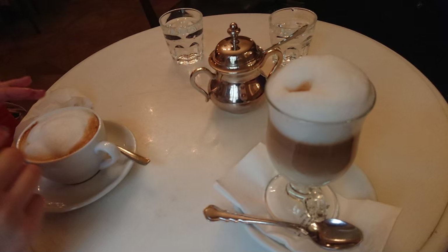 Bàn với cốc cappuccino và cốc espresso có lớp bọt sữa ấm ấm ấm và bình trà bạc cùng với ly trong suốt