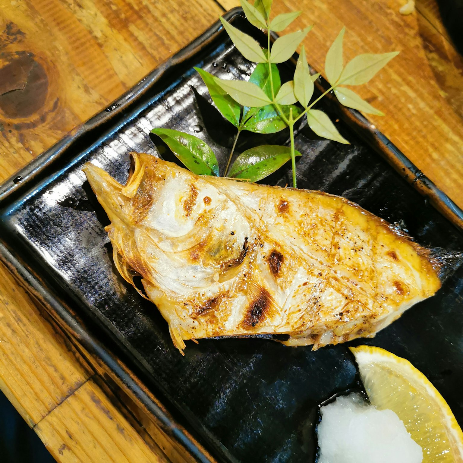 Hidangan ikan bakar di atas piring hitam dengan lemon dan daikon parut dihiasi daun