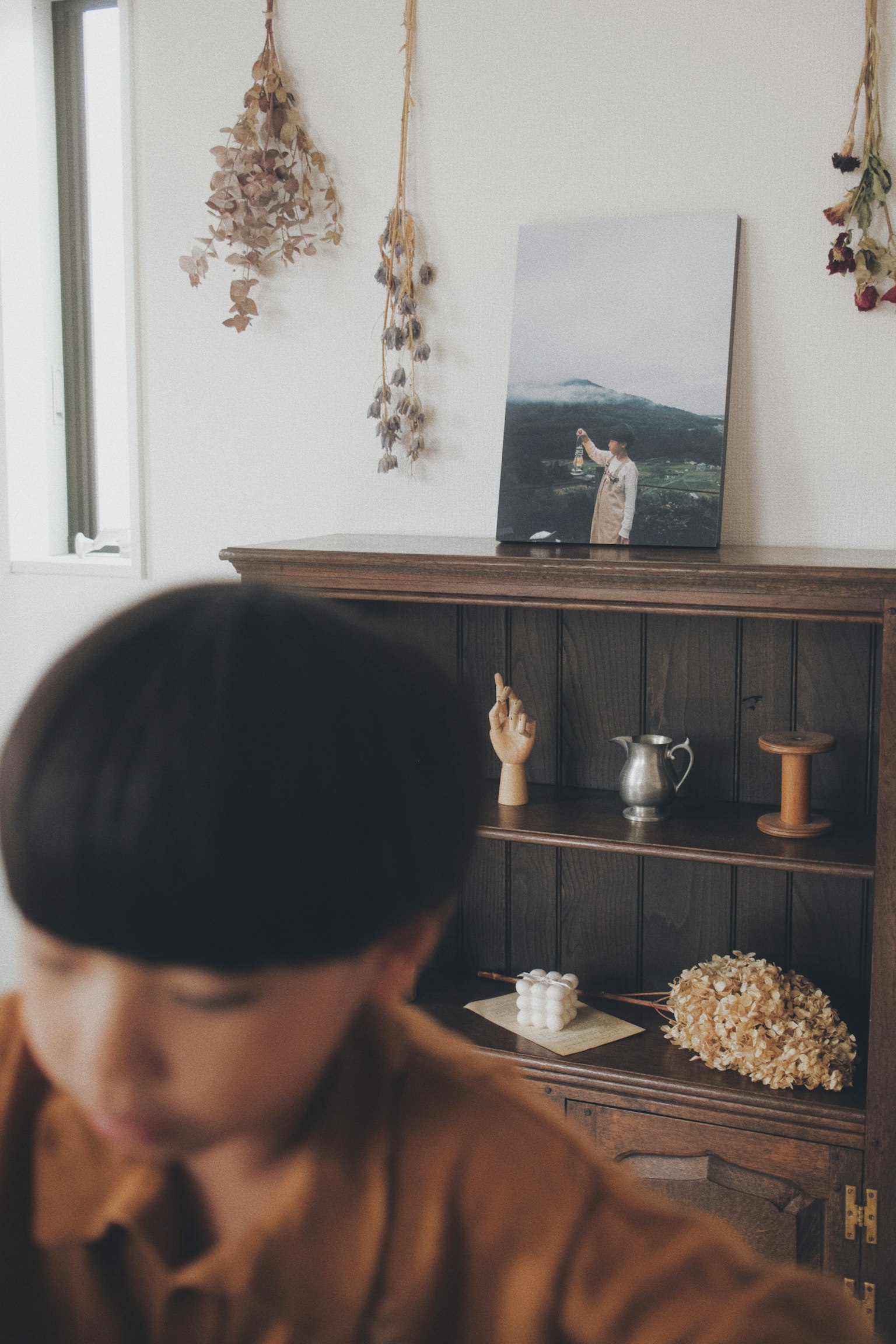 木製の棚に飾られた写真とドライフラワーがある部屋の一角で、手前にいる人物がぼやけている