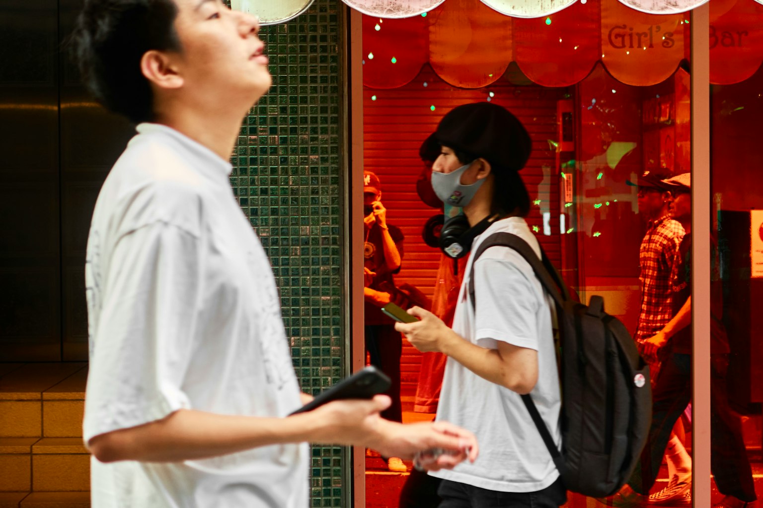 赤い背景の店の前で立っている二人の若者 一人は上を見上げている 一人はスマートフォンを見ている