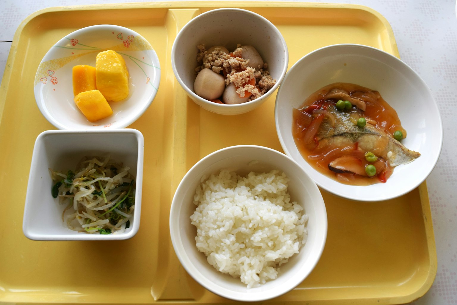 Japanese meal set on a yellow tray featuring rice, simmered vegetables, bean sprout salad, chicken dish, and mango dessert