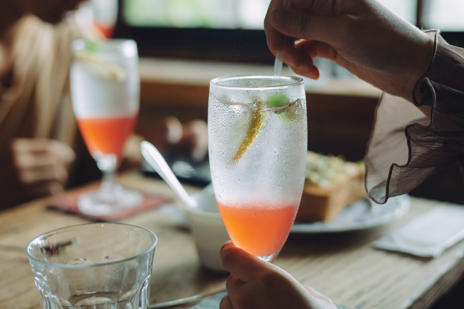 Hand holding a cocktail glass with a colorful drink and food in the background