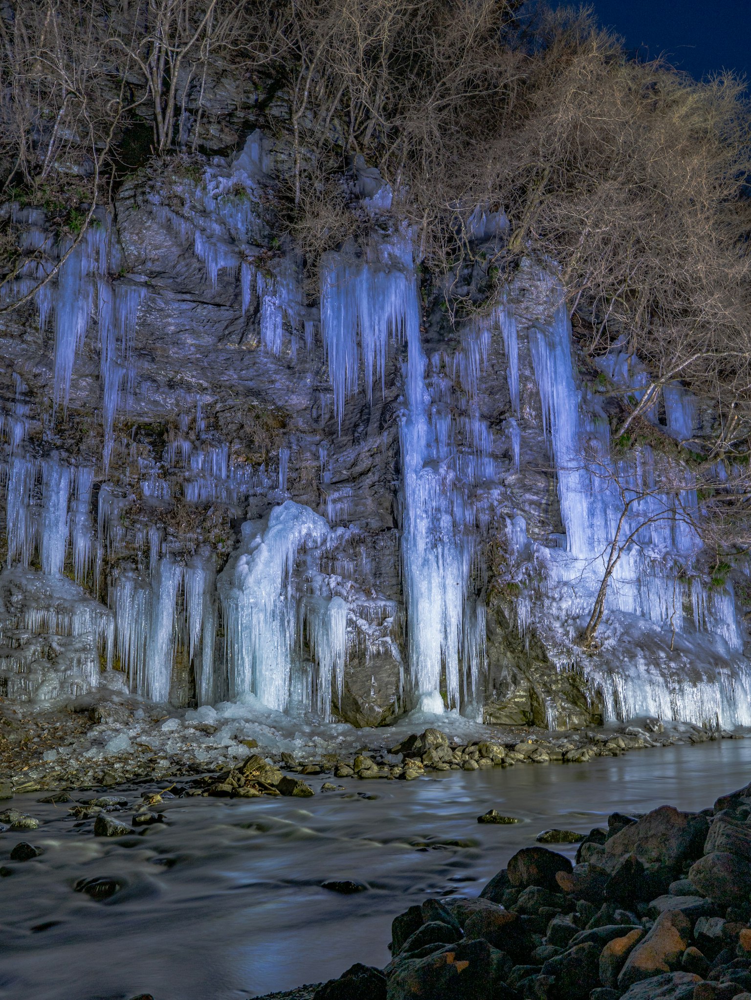 Paesaggio invernale con una cascata di ghiaccio e una scogliera rocciosa illuminata da una luce blu