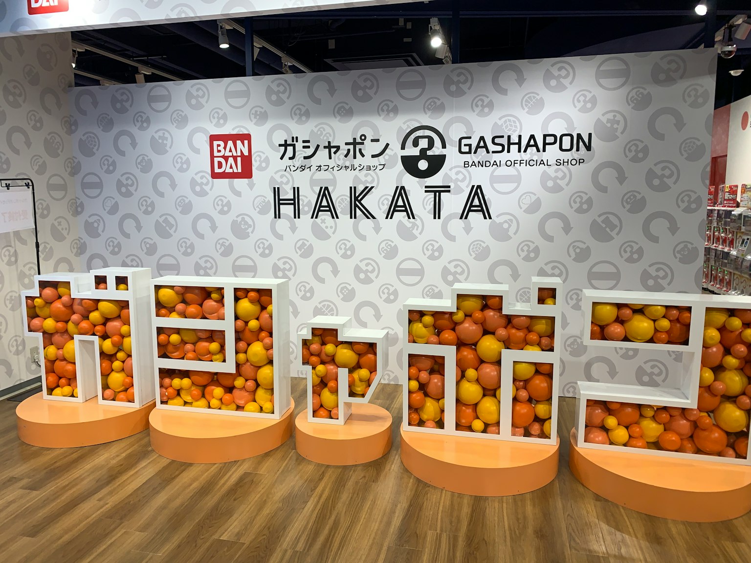 万代扭蛋展示，白色字母拼写Gashapon和Hakata，内部装满橙色球