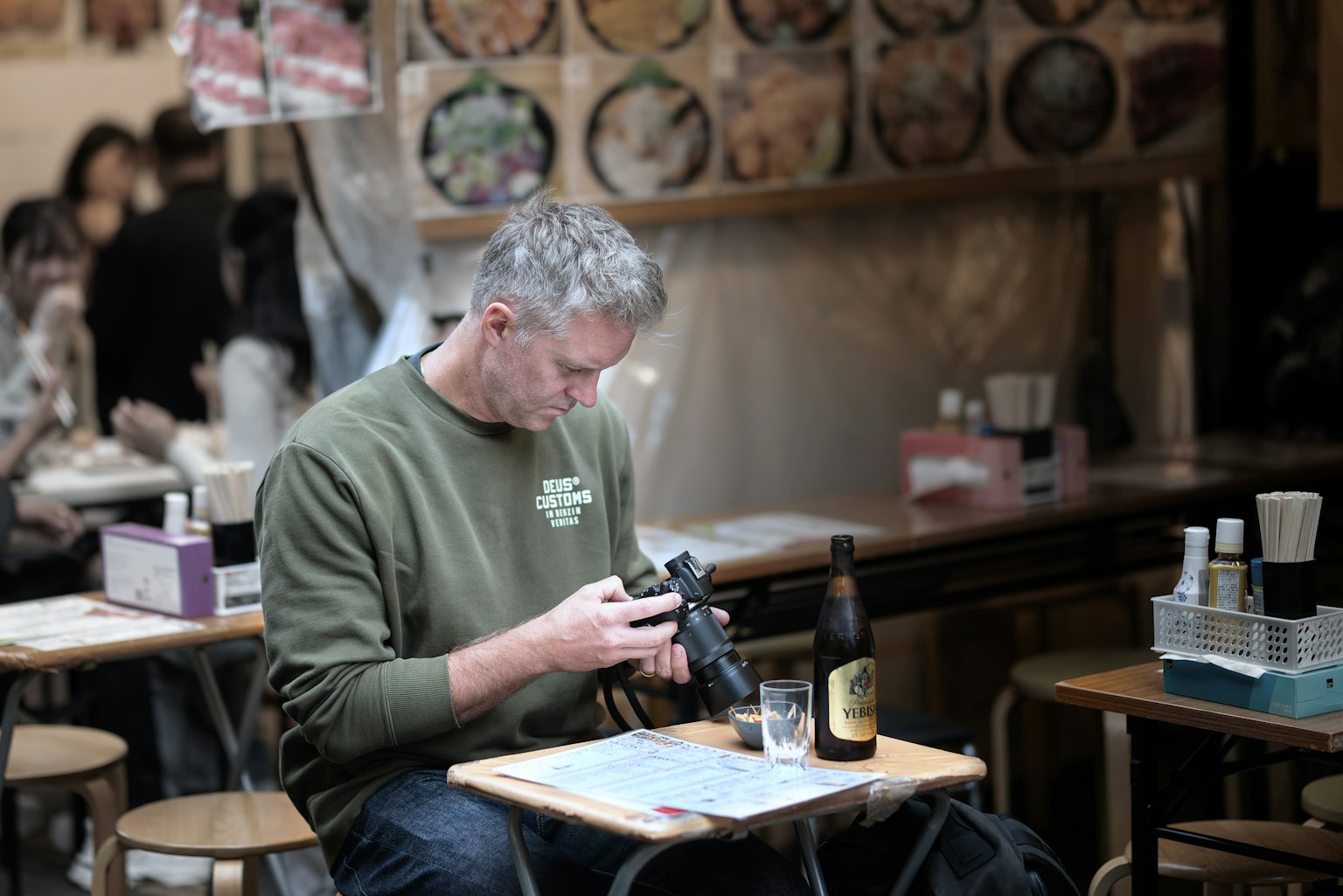 A man sitting at a table in a restaurant holding a camera and a bottle of beer