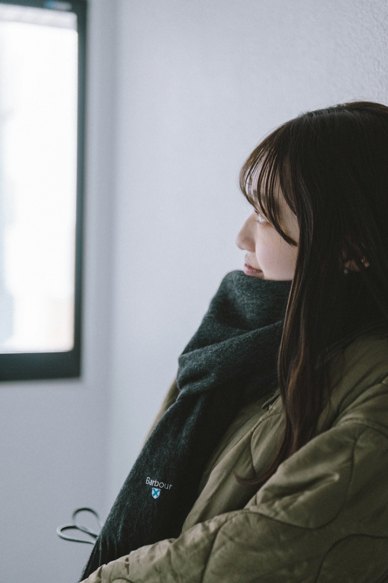 A woman looking sideways by a window wearing a warm coat and scarf