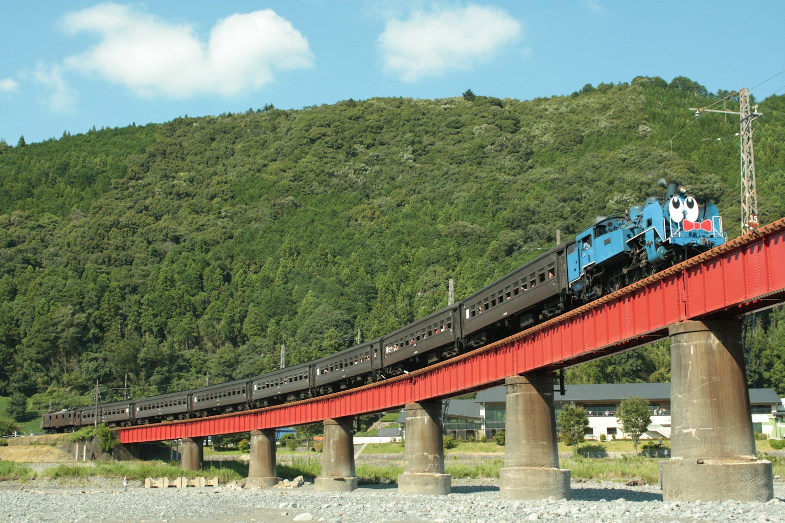 Una locomotora de vapor azul cruzando un puente rojo con montañas verdes y cielo azul de fondo