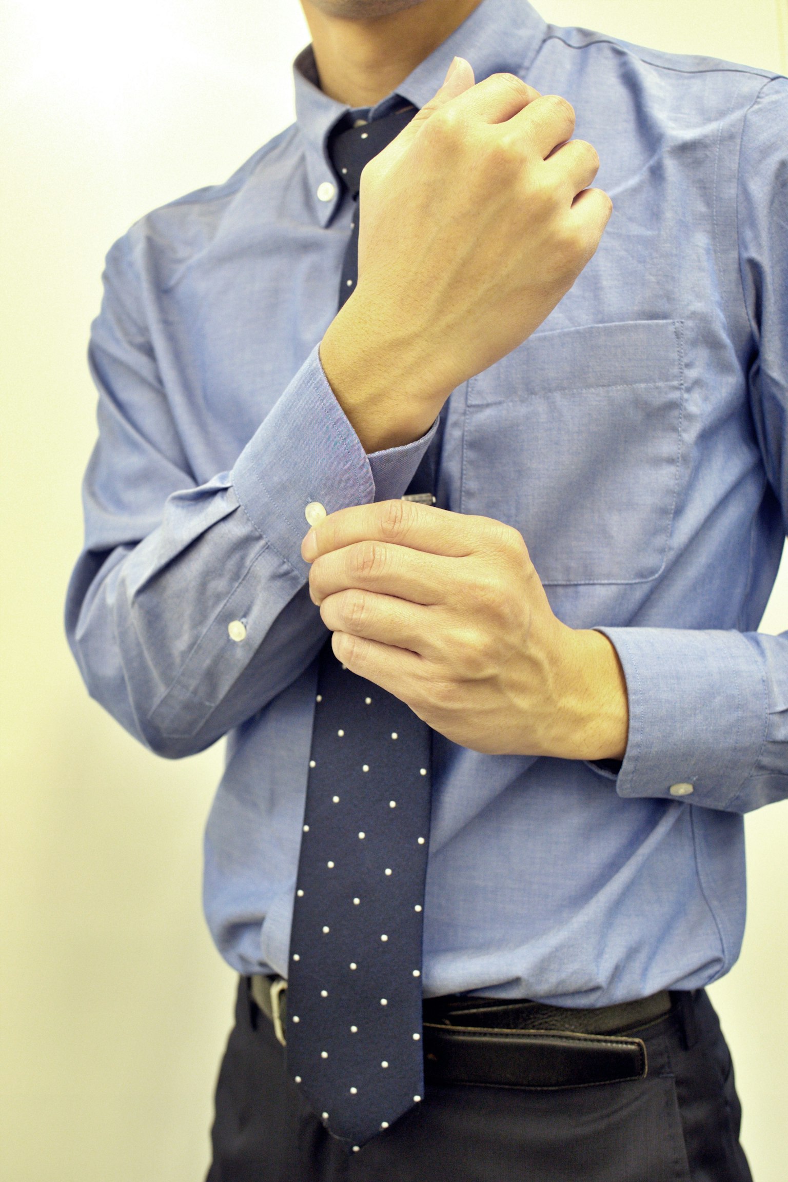 Man in a blue shirt adjusting his cuff while preparing to tie a polka dot tie